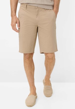 Man draagt beige casual shorts en bijpassende slip-on schoenen, staand met één hand in de zak tegen een witte achtergrond.