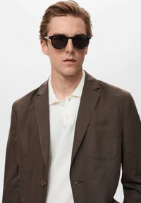 Brun blazer med en enkelt lomme, parret med en hvid tekstureret polo skjorte. Model iført sorte solbriller. Ren, moderne æstetik.