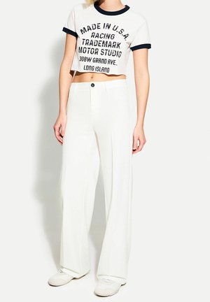 Kobieta nosząca białe spodnie o szerokich nogawkach, połączone z białym krótkim t-shirtem z czarnym napisem i czarnym wykończeniem na rękawach i kołnierzu.