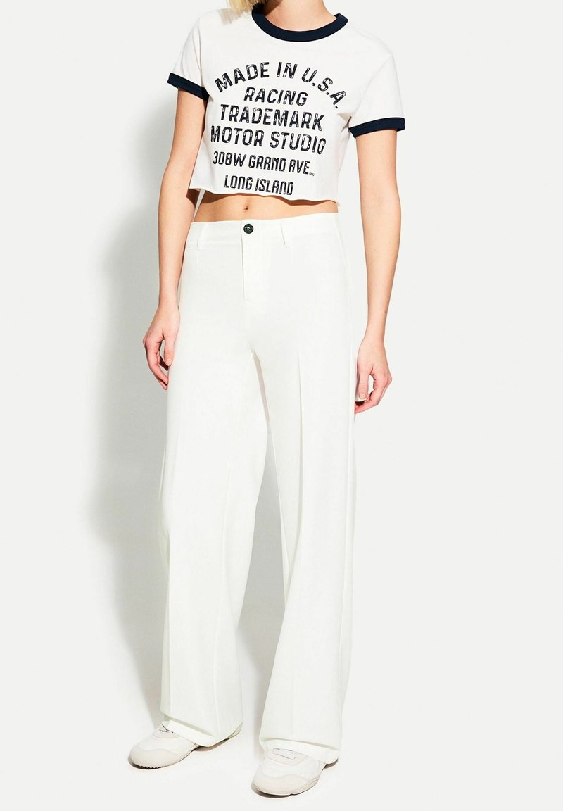 Femme portant un pantalon blanc à jambes larges associé à un t-shirt court blanc avec un texte noir et un liseré noir aux manches et au col.
