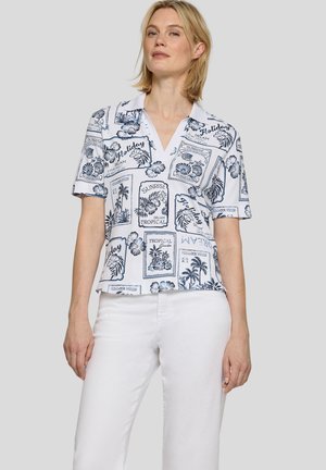 Blonde Frau trägt ein weißes kurzärmeliges Hemd mit blauen tropischen und ferienbezogenen Motiven und weiße Hosen vor schlichtem Hintergrund.