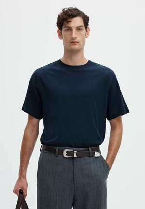 Ung mand med mørkt krøllet hår iført en navyblå t-shirt, grå bukser og et mørkt bælte, der holder en brun taske i den ene hånd.