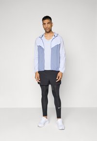 Világoskék és fehér zipzáros dzseki, két elülső zsebbel, fehér ing felett viselve, fekete rövidnadrággal és fekete leggings-szel; fehér sportcipők.