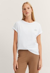Weißes Baumwoll-T-Shirt mit umgeschlagenen Ärmeln und lockerer Passform. Besitzt einen kleinen Druck auf der Brust. Getragen mit braunen Hosen.
