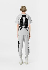 Camiseta de algodón gris con gráficos de alas negras en la parte posterior, combinada con pantalones de chándal grises a juego que presentan acentos gráficos y detalles en los bolsillos.