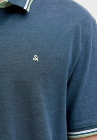 Polo shirt in donkerblauwe stof met een gestructureerd patroon. Heeft een bicolor kraag met groene en witte strepen en een klein wit logo.