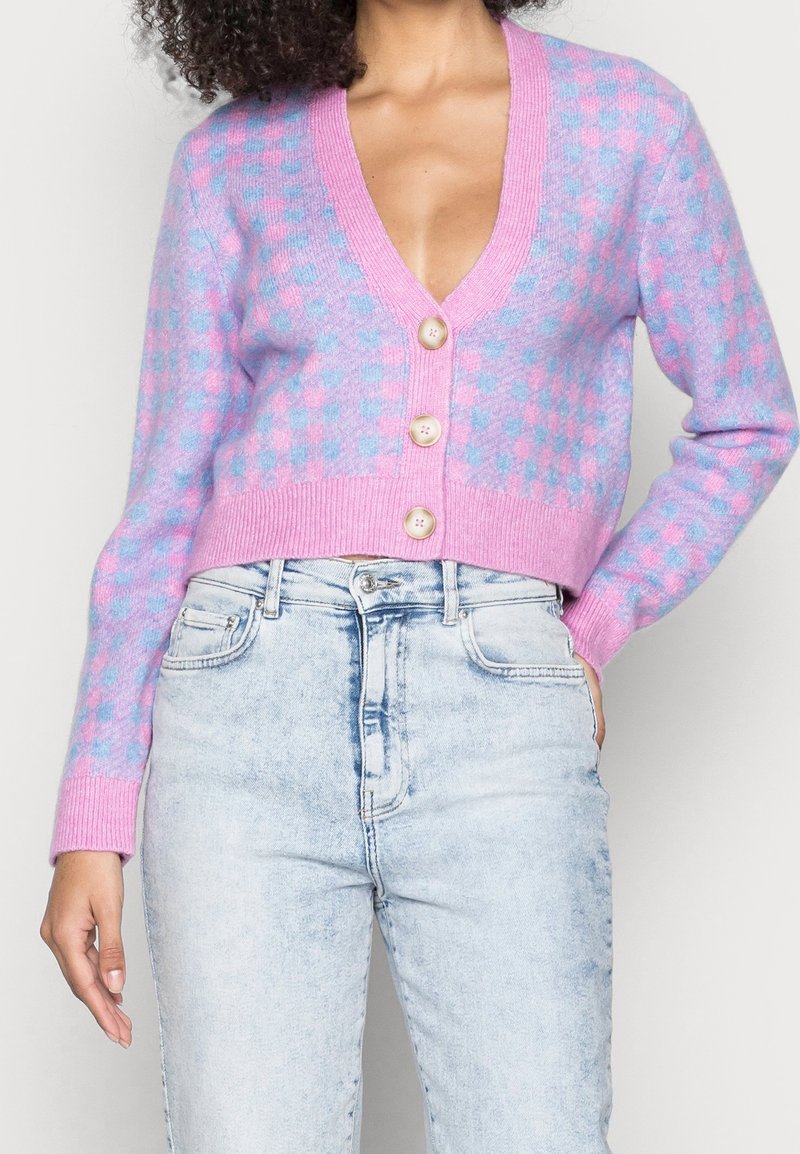 Femme portant un cardigan court à carreaux rose et bleu avec des boutons beige et un jean taille haute délavé clair, main posée dans la poche.