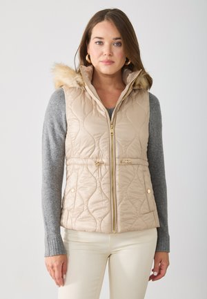 Beige quilte vest med falsk pelshette, gullglidelås og innsnevret midje. Båret over en grå genser, med kremfargede bukser.