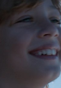Gros plan sur le visage d'un enfant avec des cheveux châtain clair, des yeux bleus et une expression souriante. Texture de peau douce, la lumière naturelle met en valeur les traits.