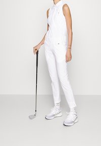 Vitt ärmlöst golfset med krage, byxor med elastisk midja och vita-och-grå golfskor, som håller en golfklubba.