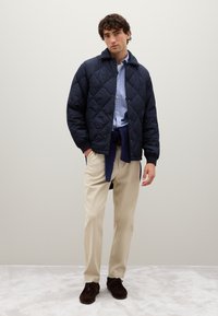 Giacca trapuntata blu navy con colletto a punta, abbinata a una camicia a righe azzurro chiaro e pantaloni beige. Modello che indossa scarpe marroni scure.