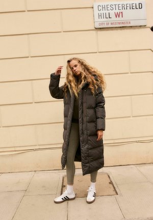 Manteau long en duvet gris avec capuche, présentant un design matelassé et des boutons sur le devant. Assorti à des leggings gris et des baskets blanches avec des rayures noires.