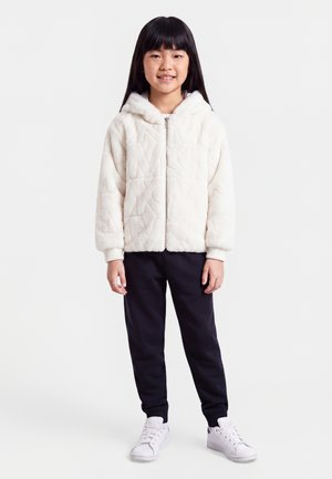 Ung pige står, iført en hvid fluffy hættetrøje med lynlås, sorte bukser og hvide sneakers, smilende med langt sort hår ned.