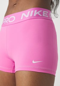 Różowe spodenki Nike Pro z szerokim elastycznym paskiem w talii, na którym znajduje się biały napis marki oraz małe białe logo. Gładka, elastyczna tkanina.