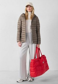 Giacca imbottita marrone, maglione bianco, pantaloni larghi grigio chiaro e sneakers argento. Tiene una borsa tote imbottita rossa brillante con un logo.