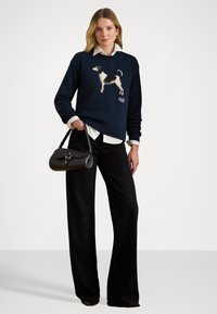 Vrouw in een marineblauwe trui met hondenprint, witte blouse, zwarte wijde broek, met een zwarte handtas aan haar zijde.