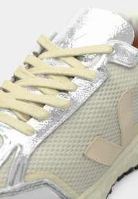 Białe siateczkowe sneakersy z srebrnymi akcentami, kremowymi sznurówkami i teksturowaną podeszwą. Wśród cech znajdują się gładka, błyszcząca powłoka i beżowe detale.