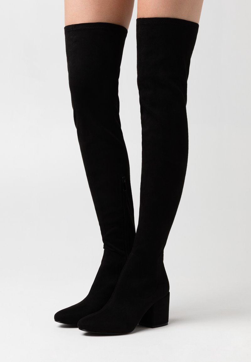 Des jambes portant des bottes en daim noires au-dessus du genou avec des talons blocs, debout sur un fond clair uni.