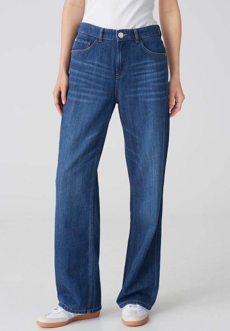 Jeans en denim bleu clair à taille haute, à jambe large, avec cinq poches et des coutures contrastées, associés à des baskets blanches.