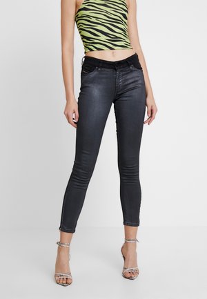 Kvinde iført sorte skinny jeans med en blank finish, grøn og sort zebra-stribet cropped top og sølvfarvede remsandaletter med høj hæl.