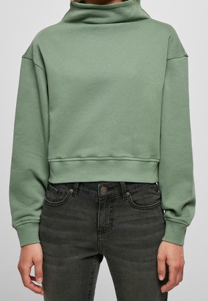 Mujer con sudadera corta de color verde salvia, con un alto cuello doblado y mangas largas, combinada con vaqueros negros de talle alto.