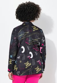 Blazer noir avec des motifs vibrants de style graffiti en rose, jaune, bleu et blanc ; design structuré avec une fermeture à bouton unique à l'avant.