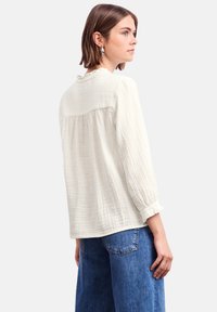 Top bianco strutturato con maniche lunghe, schiena arricciata e scollatura a volant, abbinato a jeans blu a gamba larga.