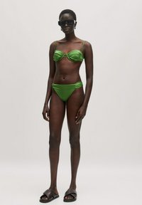 Groene twee-delige bikini met geplooide details, dunne bandjes en een lage taille; model draagt een oversized zwarte zonnebril en bruine sandalen.