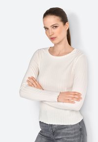 Maglietta bianca a maniche lunghe con scollo rotondo, caratterizzata da polsini e orlo sfrangiati, abbinata a jeans di denim grigi.