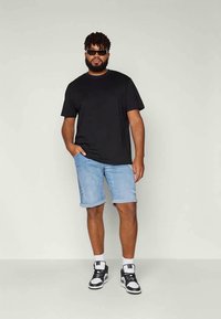 Man standing wearing black t-shirt, light blue rolled-up denim shorts, black and white sneakers, sunglasses, with one hand in pocket.