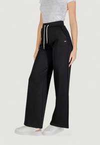 Pantalones de chándal anchos y negros con un patrón texturizado, cinturilla elástica y cordones. Combinados con zapatillas blancas.