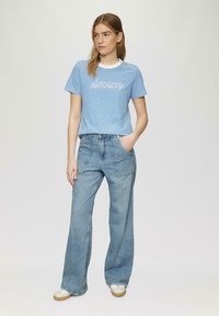 T-shirt cropped rayé bleu avec le texte « rêve » en blanc, associé à un jean large bleu clair. Le mannequin se tient avec une main dans la poche, portant des chaussures neutres.