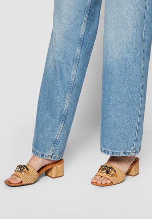 Pieds en jean droit bleu clair portant des mules à talons blocs tressés couleur cuir clair avec un large détail de chaîne métallique à l'avant.