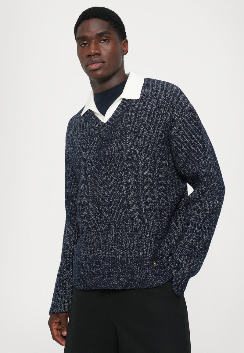 Mann in einem navyblauen strukturierten Strickpullover über einem weißen Hemd mit Kragen und schwarzen Hosen, der vor einem schlichten weißen Hintergrund steht.