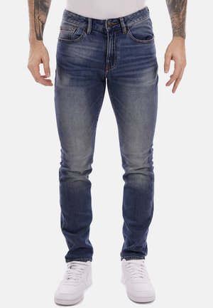 Jeans da uomo in denim blu con una vestibilità slim, lavaggio sbiadito, design a cinque tasche e chiusura con bottone, abbinati a sneakers bianche.