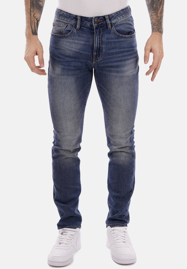 Jeans da uomo in denim blu con una vestibilità slim, lavaggio sbiadito, design a cinque tasche e chiusura con bottone, abbinati a sneakers bianche.