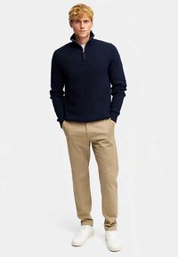 Marineblauwe gebreide trui met kwartritssluiting en geribbelde textuur, gecombineerd met beige chinos en witte sneakers, wat een casual outfit vormt.