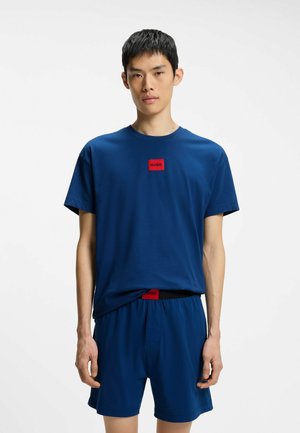 Young man wearing a blue short-sleeve shirt and matching shorts with red HUGO logos on chest and waistband, standing against a white background.