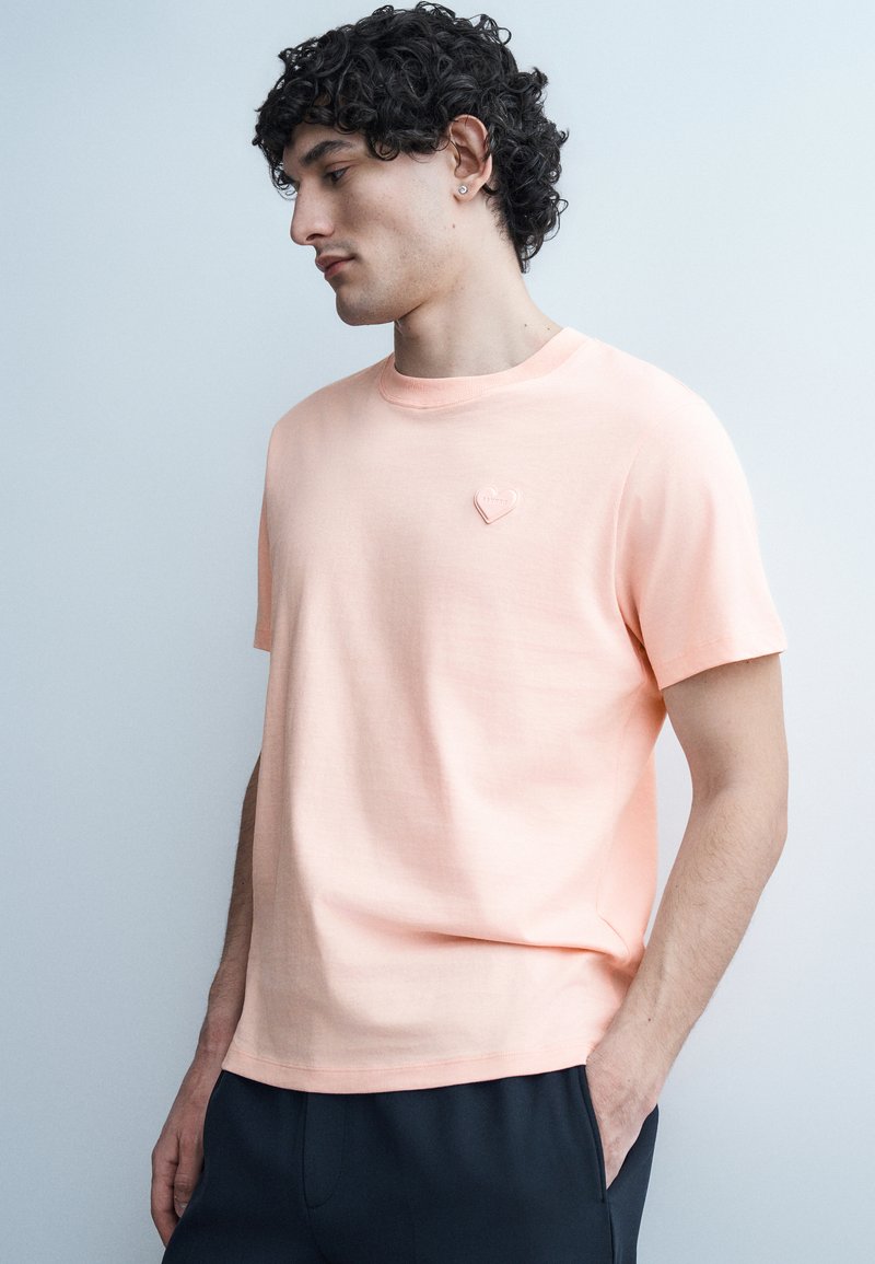 Νεαρός άνδρας με σγουρά μαλλιά που φοράει ένα ανοιχτό ροδακινί κοντομάνικο μπλουζάκι με ένα μικρό σήμα σε σχήμα καρδιάς στο στήθος, με τα χέρια στις τσέπες.