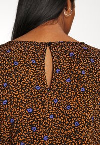 Kvinna med långt svart hår bär en svart klänning med mönster av orangea löv och små lila blommor, knappstängning är synlig vid nacken.