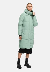 Kvinna som bär en lång mintgrön vadderad kappa med huva, svart kjol och svarta kraftiga stövlar, stående mot en enfärgad bakgrund.