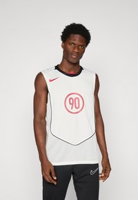 En atletisk mann iført hvit ermeløs sportstrøye med rød "90" og Nike-logo, sammen med svarte Nike-bukser, står mot en ensfarget bakgrunn.