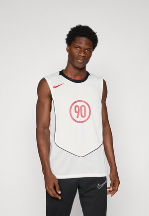 En atletisk mann iført hvit ermeløs sportstrøye med rød "90" og Nike-logo, sammen med svarte Nike-bukser, står mot en ensfarget bakgrunn.