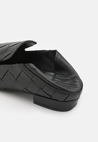 Scarpa nera in pelle slip-on con un design intrecciato, punta aperta e tacco basso di colore nero uniforme. Texture liscia con un leggero lucido.
