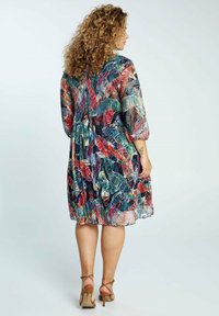 Nainen, jolla on kiharat hiukset, pukeutuneena värikkääseen, polvipituiseen lehtikuvioiseen mekkoon ja ruskeisiin korkokenkiin, seisoo selin.