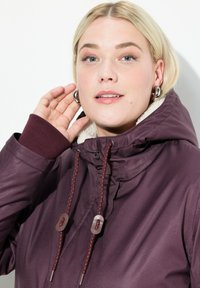 Maroonová bunda s kapucí, která má měkkou bílou podšívku, elastické manžety a dekorativní šňůrky. Ruka modelu je položena vedle obličeje.