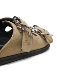 Gros plan d'une chaussure en daim beige avec une semelle noire, dotée de deux grandes boucles métalliques argentées.