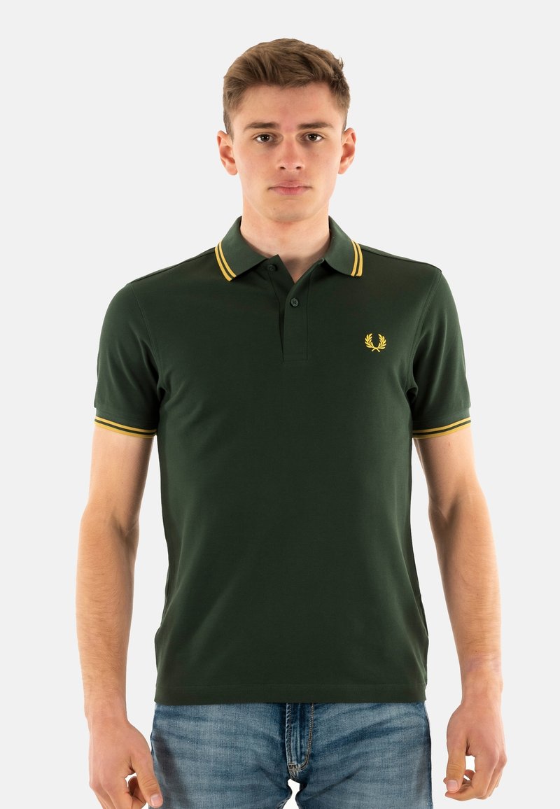 Donkergroene polo met een gele gestreepte kraag en manchetten. Heeft een klein geel embleem op de linkerborst. Korte mouwen, slim fit.