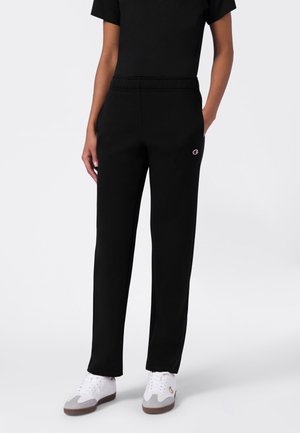 Personne portant un pantalon de survêtement noir à jambe droite avec un petit logo sur la cuisse, associé à des baskets blanches et grises, debout devant un fond uni.