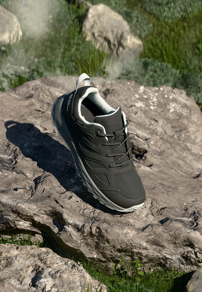 Sapatilha atlética preta com parte superior sintética lisa, painéis laterais texturizados, sola cinza e design com atacadores. Colocada sobre uma superfície rochosa com vegetação.